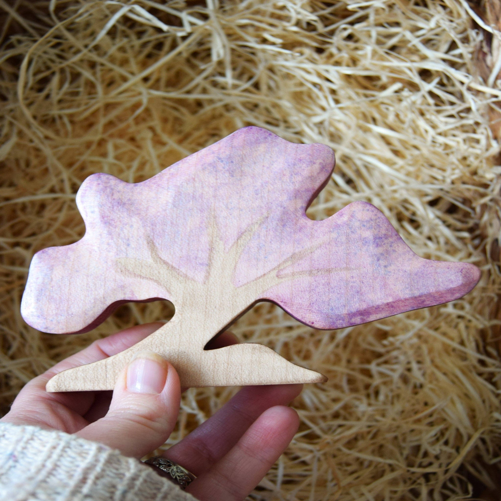 Hand holding a wooden tree with a pink and purple gradient against a straw background