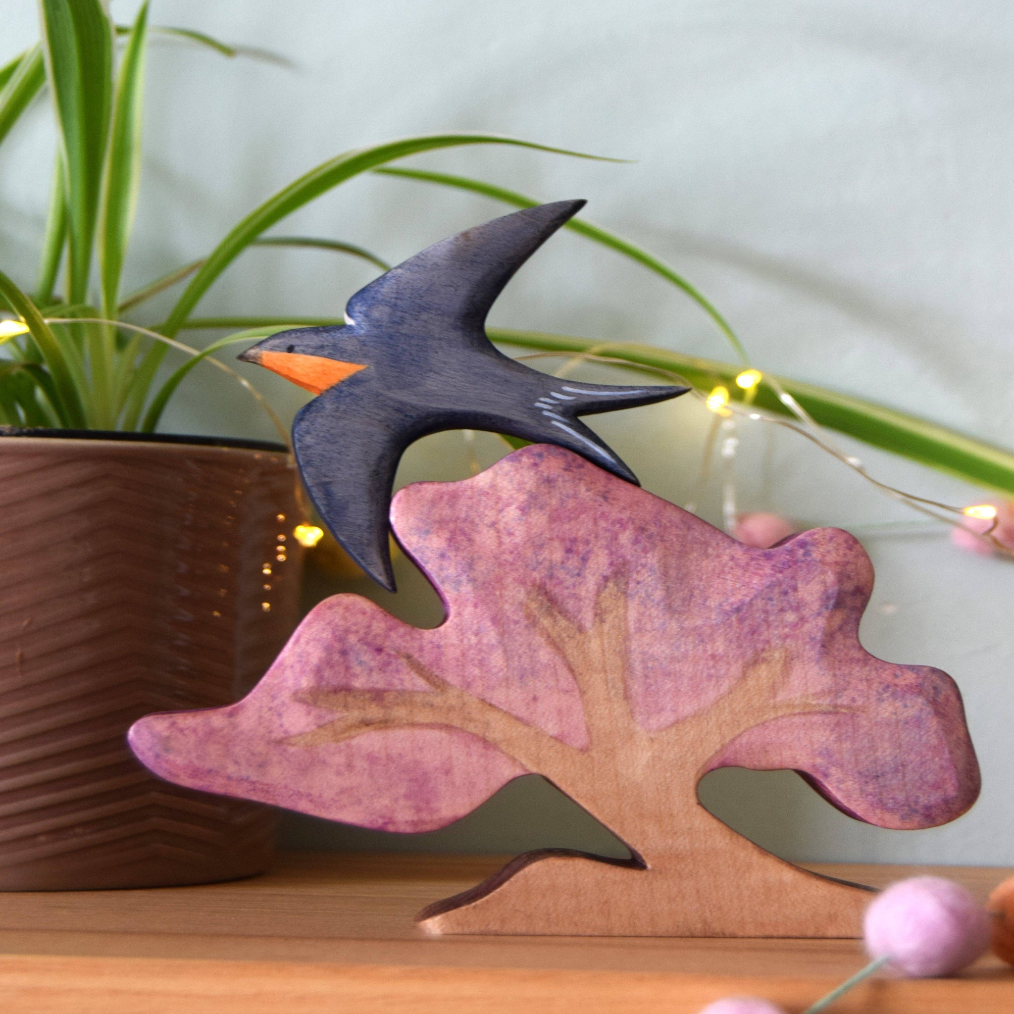 Decorative wooden leaf with a bird on top, placed on a surface with a plant and lights in the background.