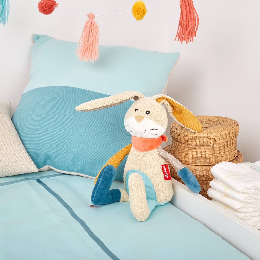 Plush bunny toy with a red tag on a bed with blue bedding and colorful tassels in the background.