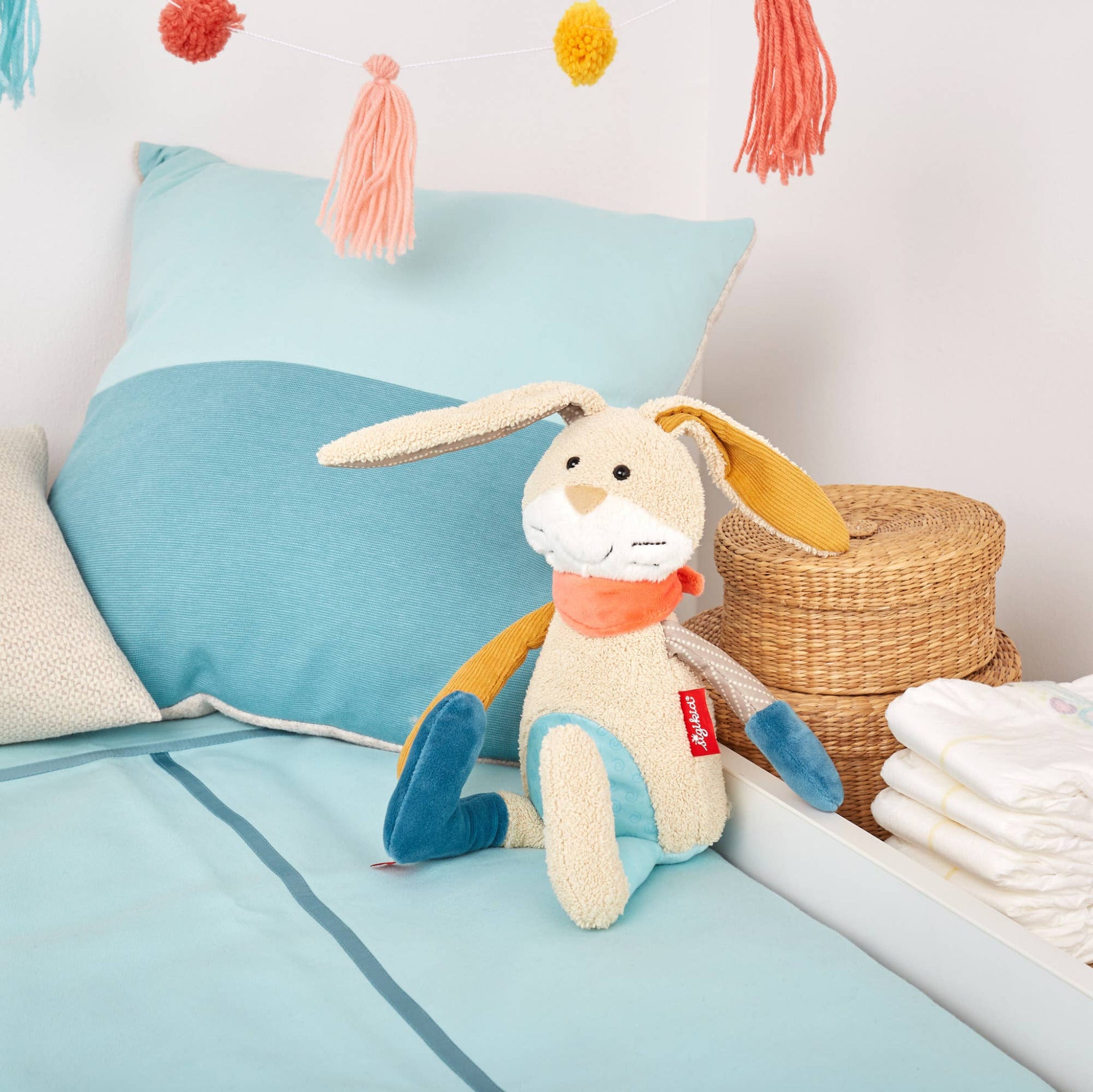 Plush bunny toy with a red tag on a bed with blue bedding and colorful tassels in the background.