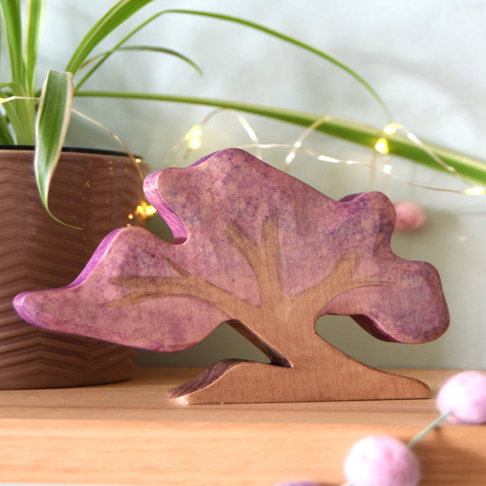 Wooden tree-shaped object with a purple finish on a wooden surface with a plant and lights in the background.